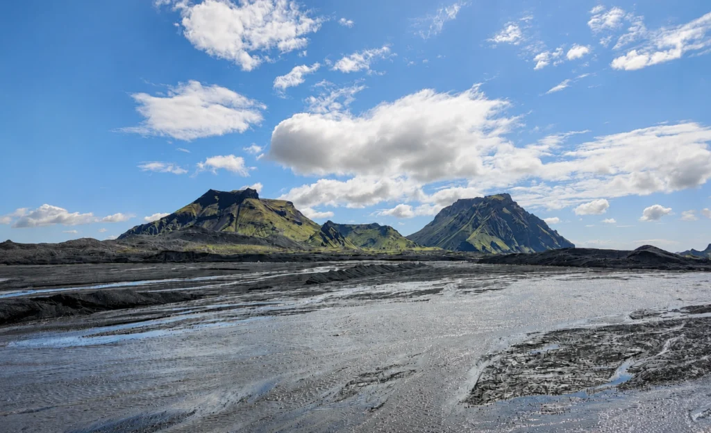 Paisaje volcánico en Islandia con llanura de ceniza y montañas verdes bajo cielo azul
