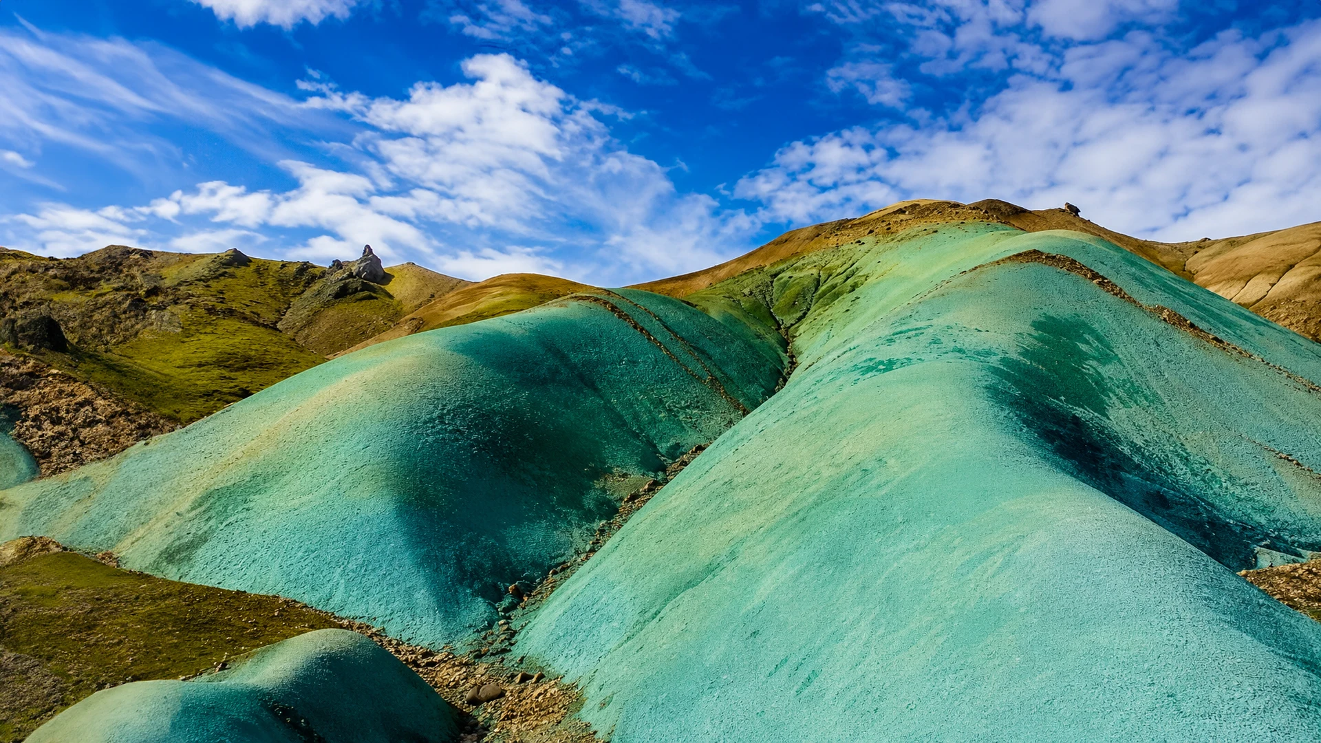 Primer plano de colinas turquesas y verdes en Grænihryggur, Islandia, bajo un cielo azul con nubes.