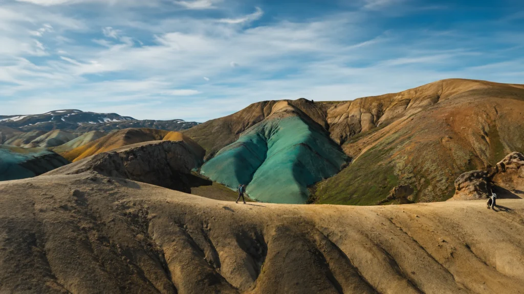 Senderistas recorren un sendero frente a una colina turquesa entre montañas ocres de Grænihryggur, Islandia.