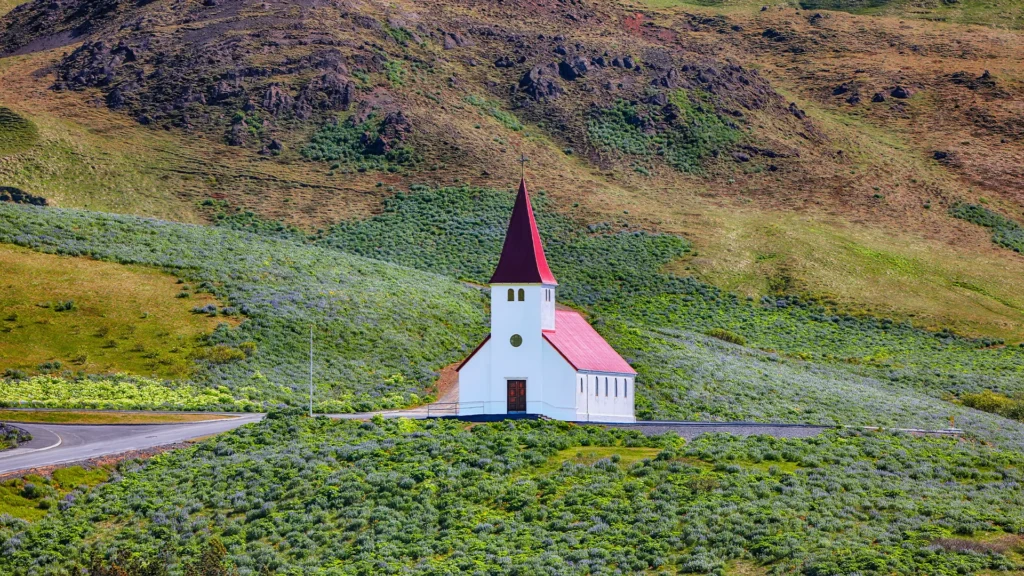Vista frontal de la iglesia Víkurkirkja rodeada de vegetación al pie de una ladera en Vík, Islandia.