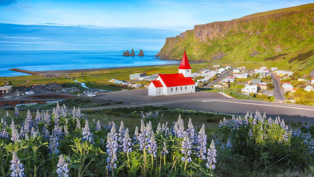Iglesia Víkurkirkja sobre Vík, con el mar, acantilados verdes y formaciones rocosas al fondo.