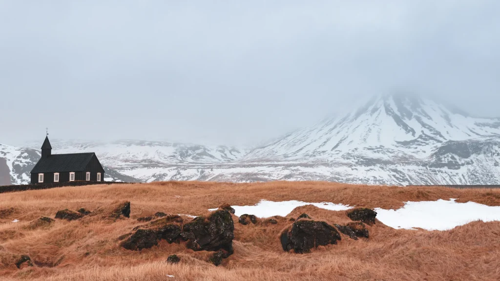 Iglesia negra en una llanura de hierba rojiza, con montañas nevadas cubiertas por la niebla al fondo.