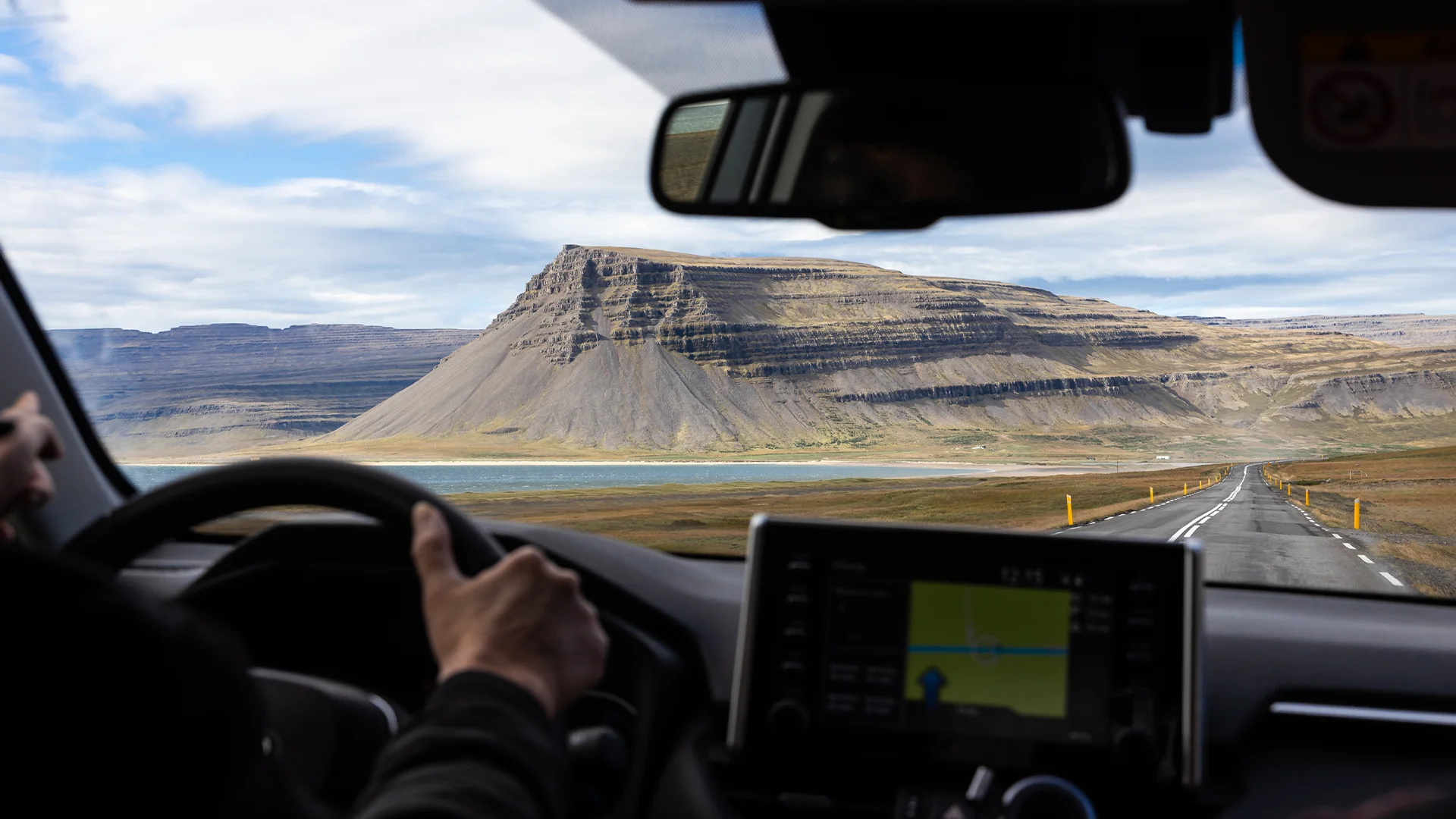 Conducción por una carretera de Islandia vista desde el interior de un coche de alquiler, con montañas y un fiordo al fondo.