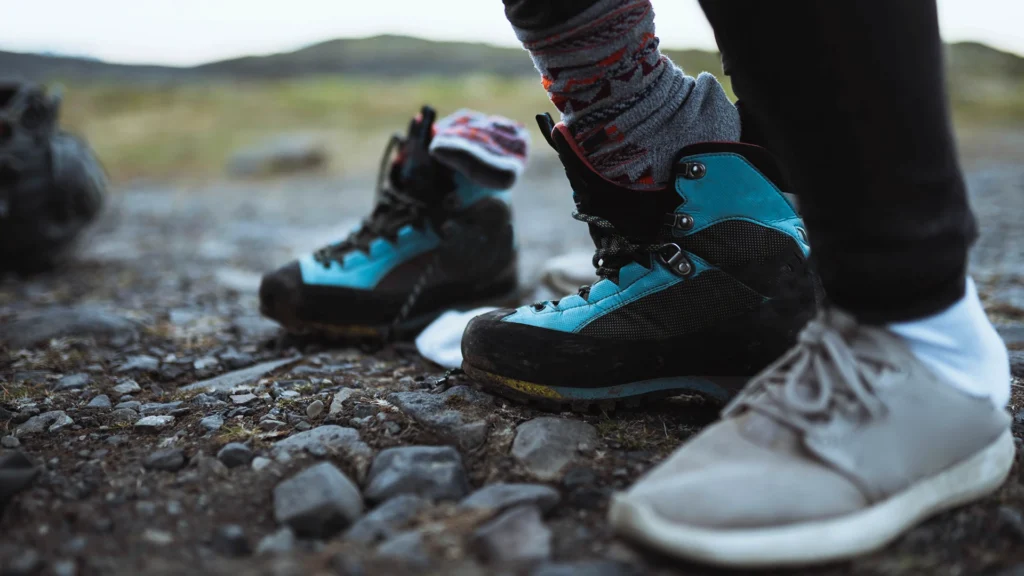 Primer plano de unas botas de senderismo y otros zapatos sobre un sendero rocoso.
