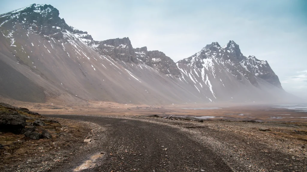 Camino de grava en Islandia con charcos y piedras, rodeado de montañas escarpadas con restos de nieve.