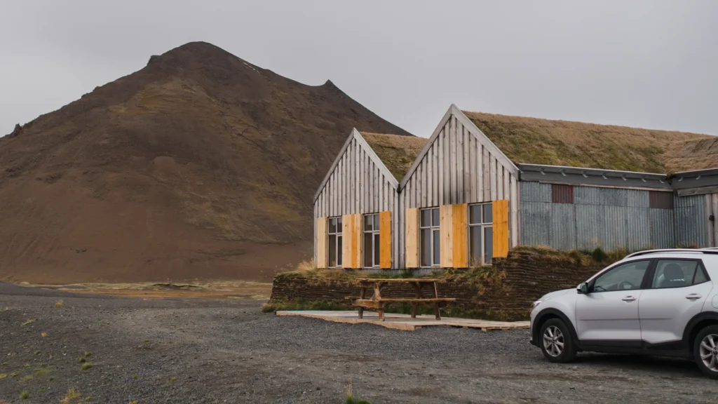 SUV blanco aparcado junto a un alojamiento rural con tejado de hierba en Islandia, rodeado de montañas y terreno volcánico.