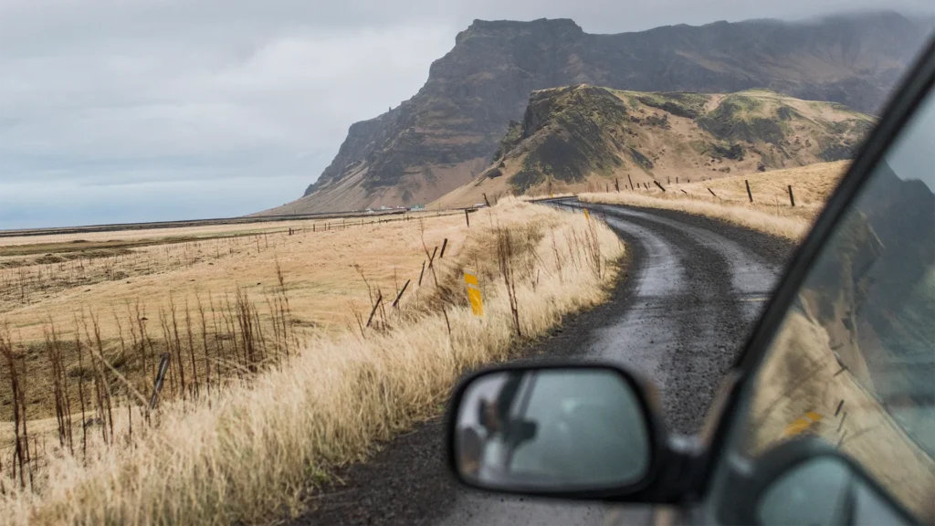 Carretera estrecha de grava mojada en Islandia, vista desde un coche, con una curva junto a montañas y campos de hierba.