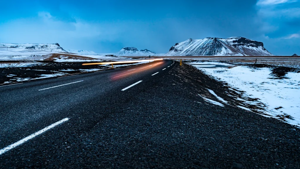 Carretera asfaltada de Islandia al anochecer, rodeada de campos nevados y montañas, con las luces de un vehículo en movimiento.