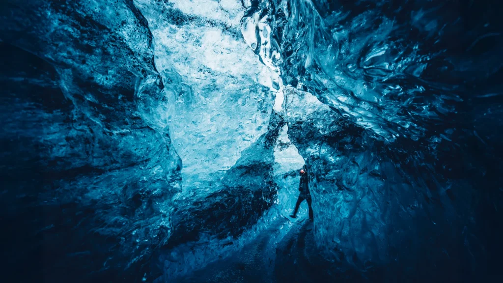 Persona caminando por el interior de una cueva de hielo azul en Islandia, rodeada de paredes glaciares con formas y texturas naturales.