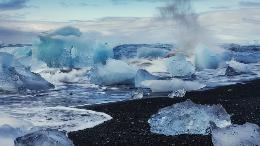 Grandes bloques de hielo azul y transparente en Diamond Beach, Islandia, mientras las olas del océano Atlántico rompen sobre la playa de arena volcánica negra.
