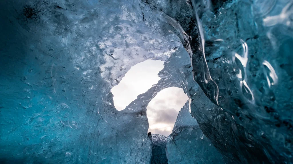 Interior de una cueva de hielo azul en Islandia, con paredes glaciares translúcidas y una abertura natural por la que entra la luz exterior.