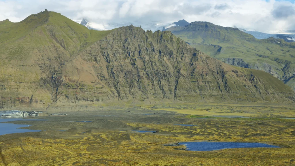 Paisaje de montañas verdes y rocosas frente a un glaciar, con charcas y llanura de sedimentos en primer plano.