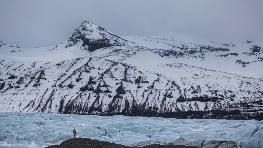 Una persona camina sobre un glaciar azul al pie de una montaña cubierta de nieve.