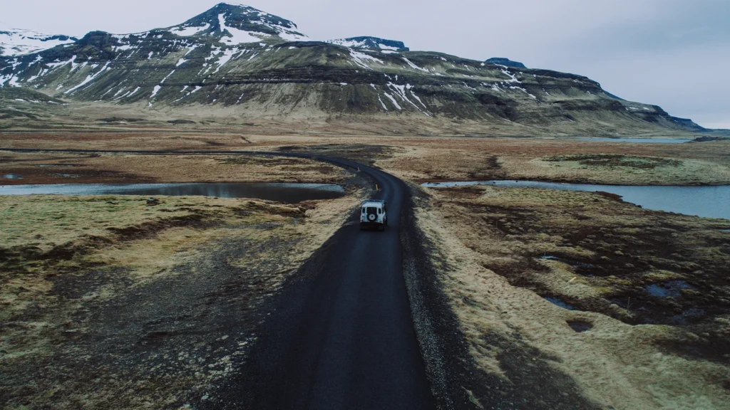 Todoterreno blanco circulando por una carretera estrecha y asfaltada de Islandia, entre lagunas y montañas con restos de nieve.