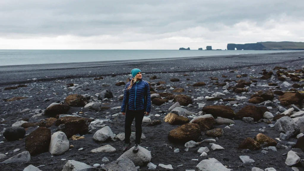 Viajera con gorro y chaqueta acolchada azul sobre una playa de arena negra y piedras.