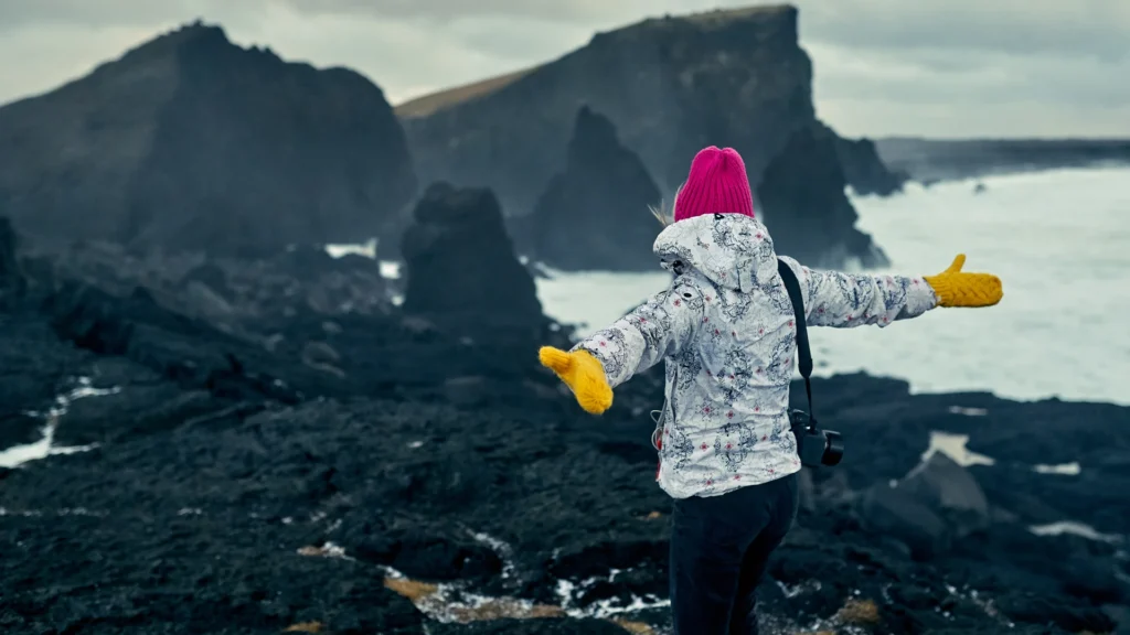 Viajera con chaqueta, gorro y guantes frente al mar, en una playa de arena negra.