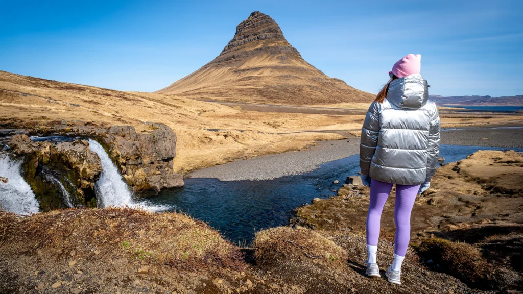 Viajera con abrigo acolchado y gorro frente a Kirkjufell y la cascada Kirkjufellsfoss.