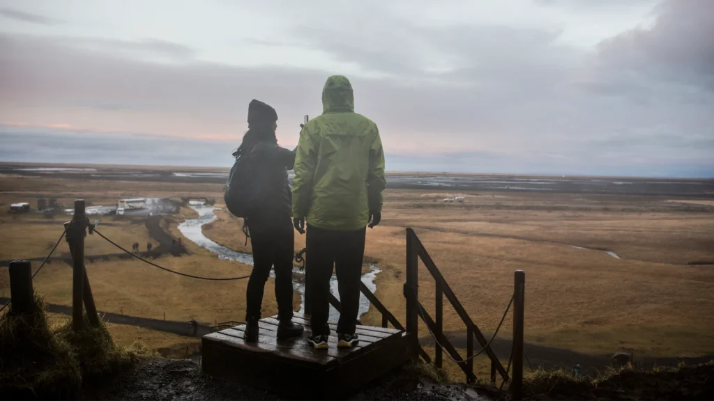 Dos viajeros con chaquetas de exterior contemplan un paisaje abierto desde un mirador.