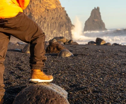 Viajero con chaqueta naranja y botas sobre una roca en la playa de arena negra de Reynisfjara, con Reynisdrangar al fondo.