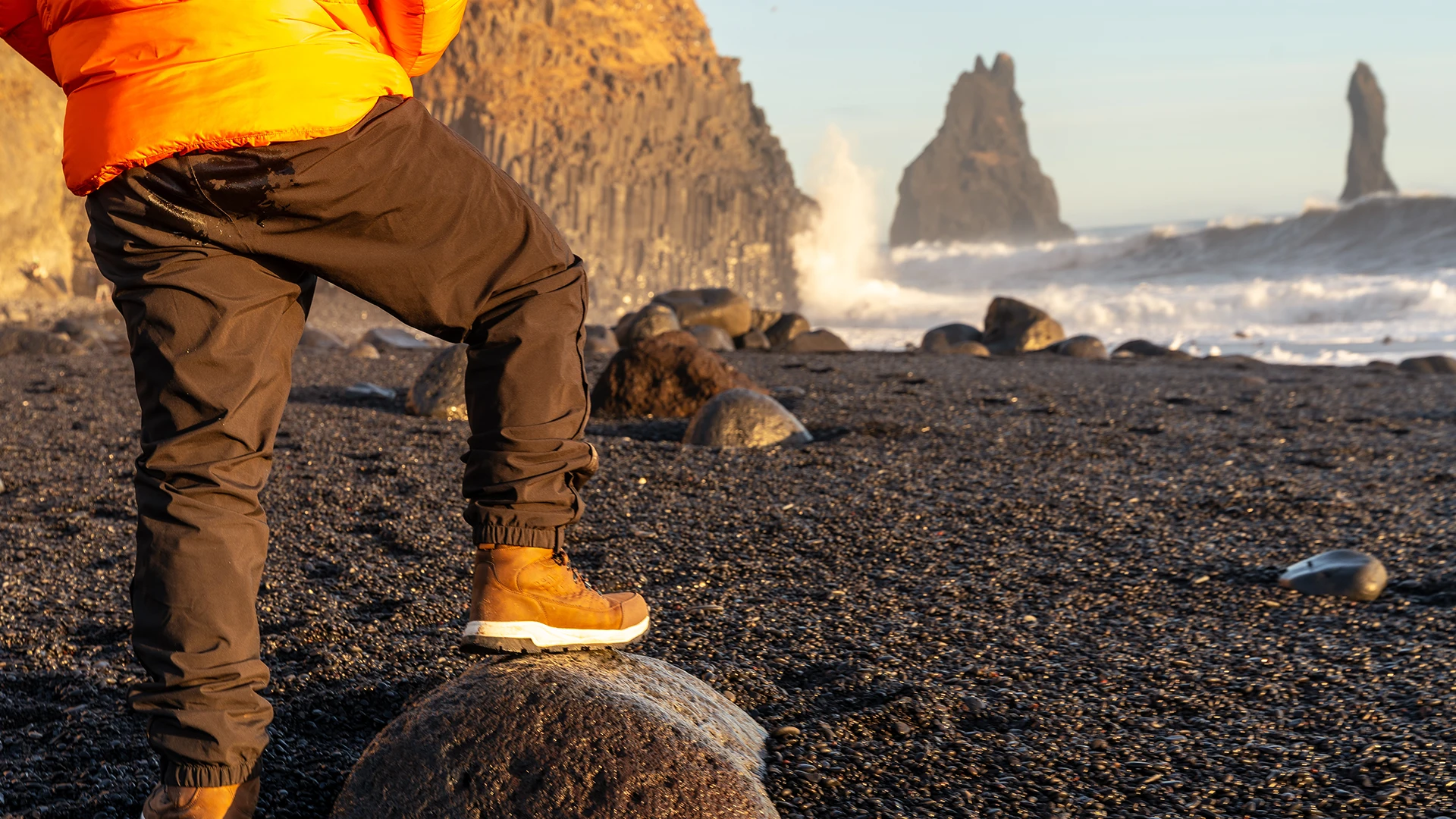 Viajero con chaqueta naranja y botas sobre una roca en la playa de arena negra de Reynisfjara, con Reynisdrangar al fondo.