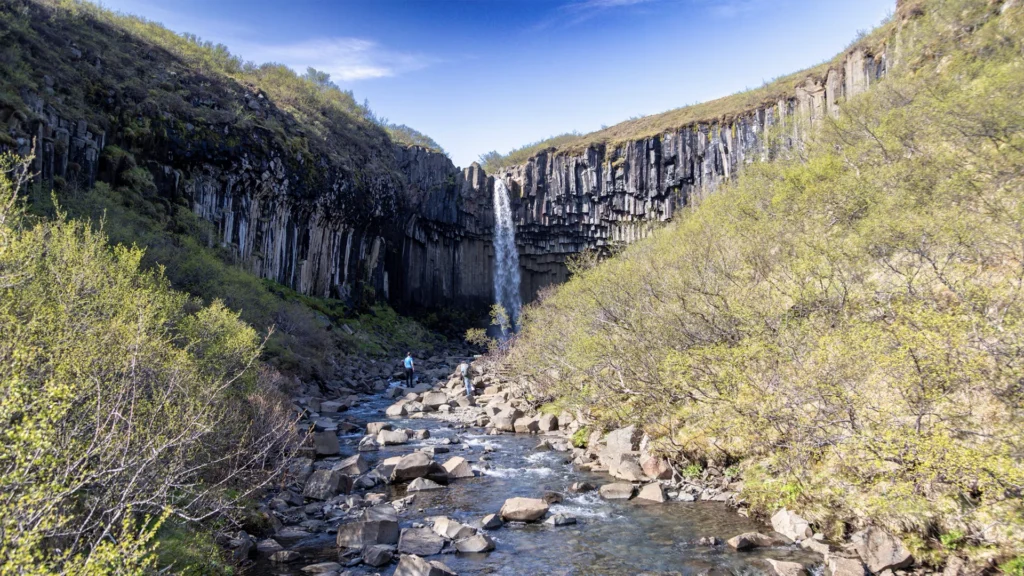 Svartifoss y sus columnas de basalto, con dos visitantes junto al arroyo en Skaftafell.